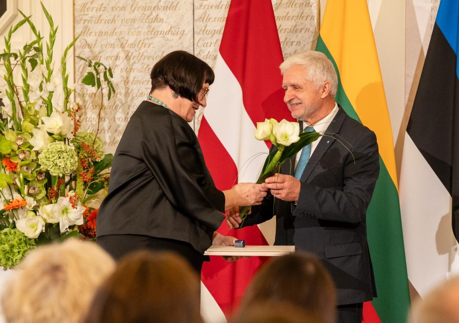 Svinīgā ceremonijā sieviete pasniedz vīrietim ziedus uz skatuves pie Latvijas, Lietuvas un Igaunijas karogiem.