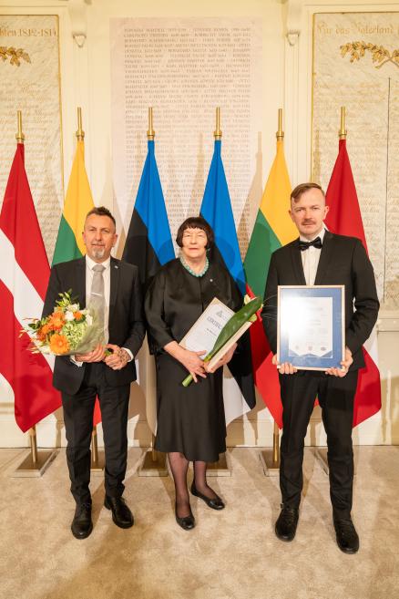 Trīs svinīgi tērpti cilvēki stāv telpā pie Latvijas, Lietuvas, Igaunijas un vēl citu valstu karogiem, turēdami ziedus un rāmī ievietotu sertifikātu.