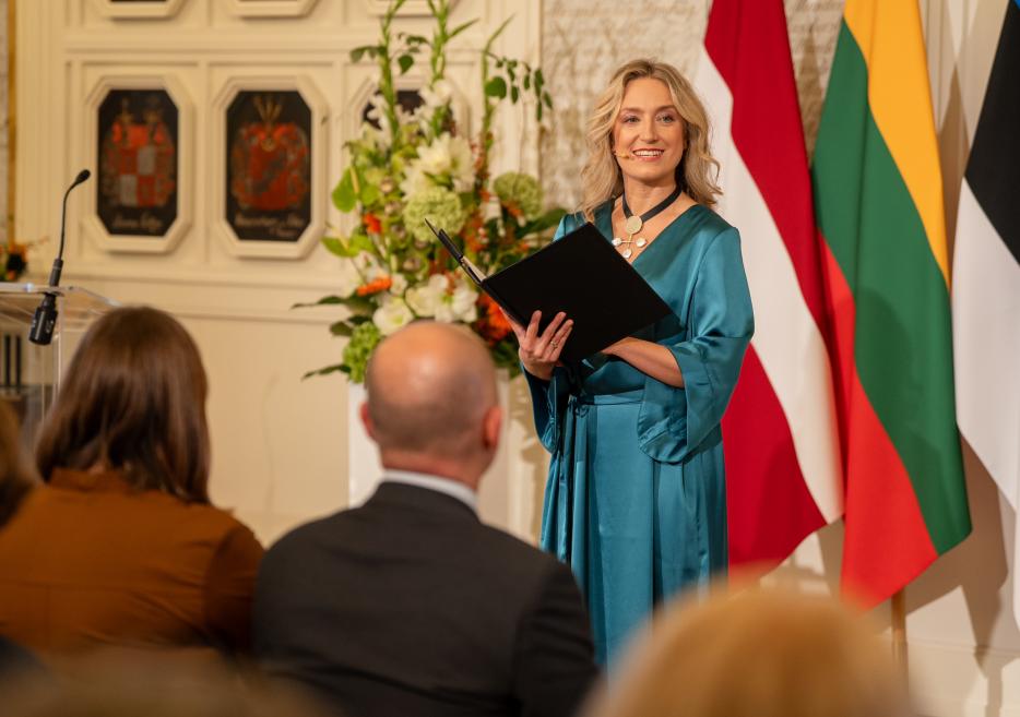 Sieviete zilā kleitā ar mapīti rokās uzstājas telpā ar Latvijas un Lietuvas karogiem fonā, kamēr priekšplānā sēž klausītāji.