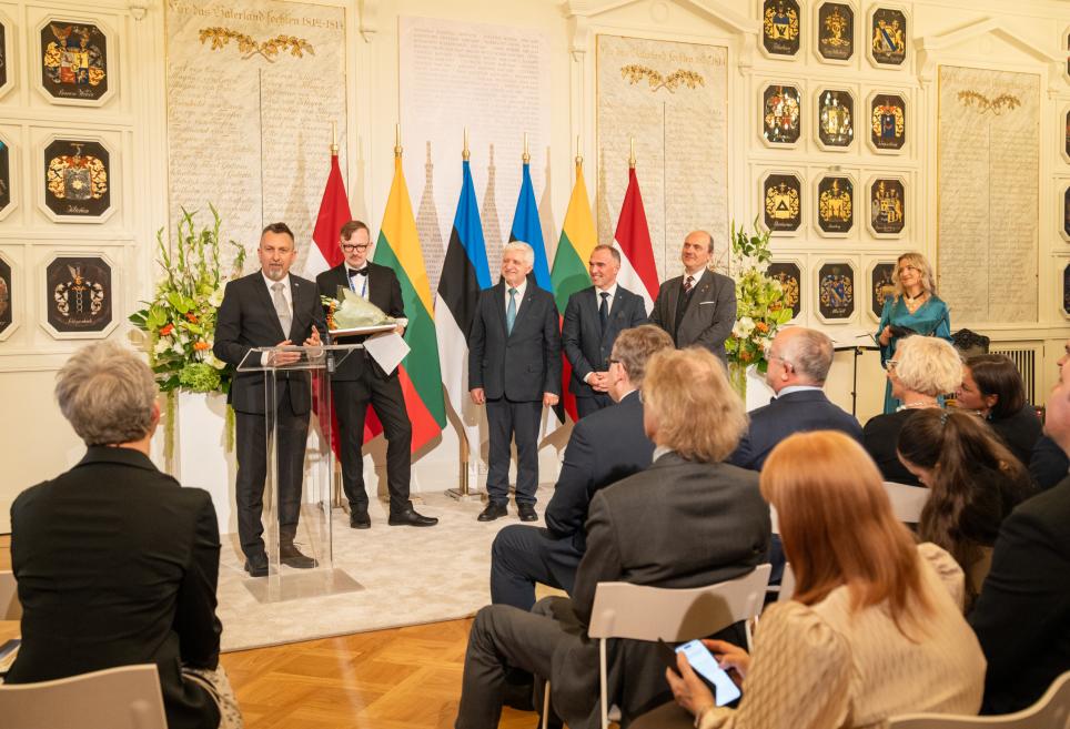 Svinīgā zālē vīrietis uzrunā sēdošu auditoriju pie stikla pults, kam fonā stāv vairāki vīrieši un sieviete starp Latvijas, Lietuvas, Igaunijas un citu valstu karogiem.