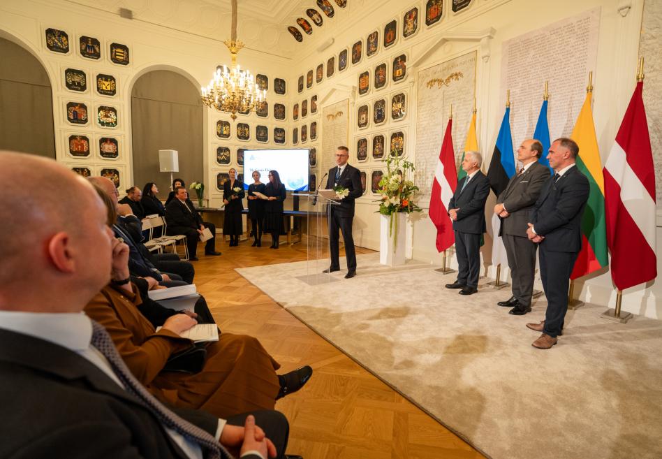 Greznā zālē ar heraldiskiem vairogiem pie sienām vīrietis runā pie pults, kamēr blakus stāv trīs vīrieši un aiz viņiem redzami Latvijas, Igaunijas, Lietuvas un Polijas karogi.