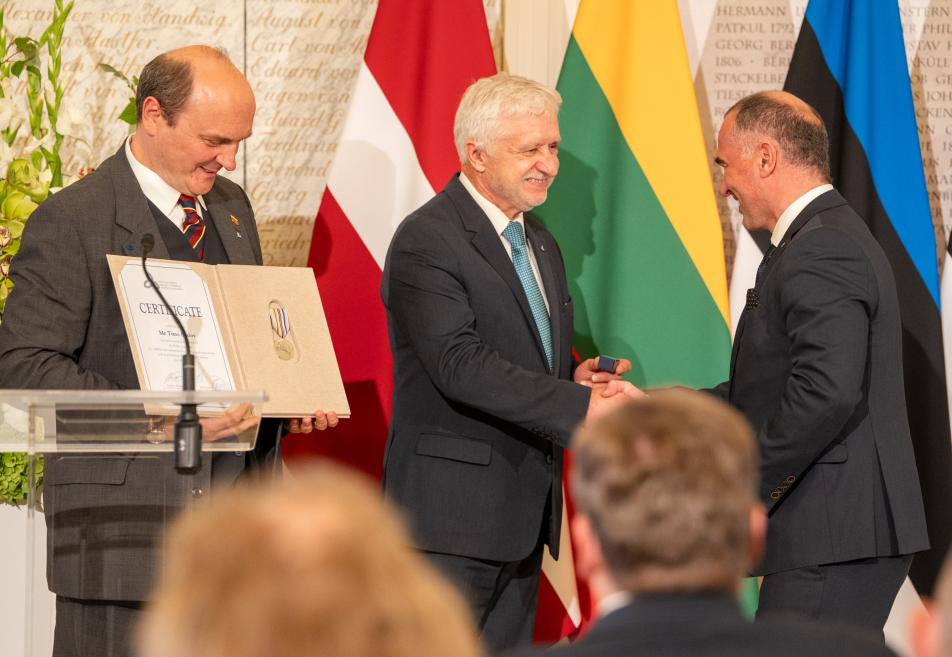 Trīs uzvalkos tērpti vīrieši oficiālā ceremonijā uz skatuves pie valstu karogiem, kur viens pasniedz sertifikātu, bet divi citi roku spiedienā apmainās ar apsveikumu.