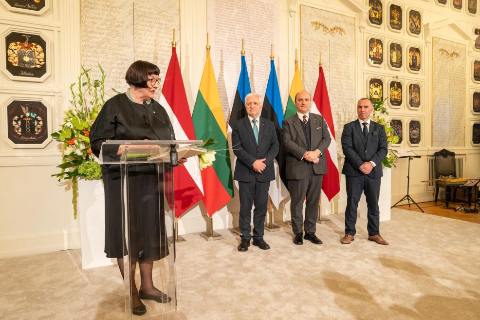 Sieviete melnā kleitā pie pults runā ceremonijas zālē, bet aiz viņas stāv trīs vīrieši uz valstu karogu fona.