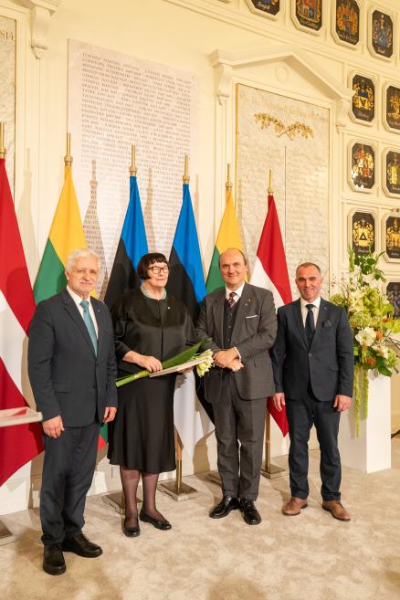 Četri uzvalkoti cilvēki stāv telpā pie valstu karogiem un sienas plāksnēm, bet vidū esošā sieviete tur baltu ziedu pušķi.