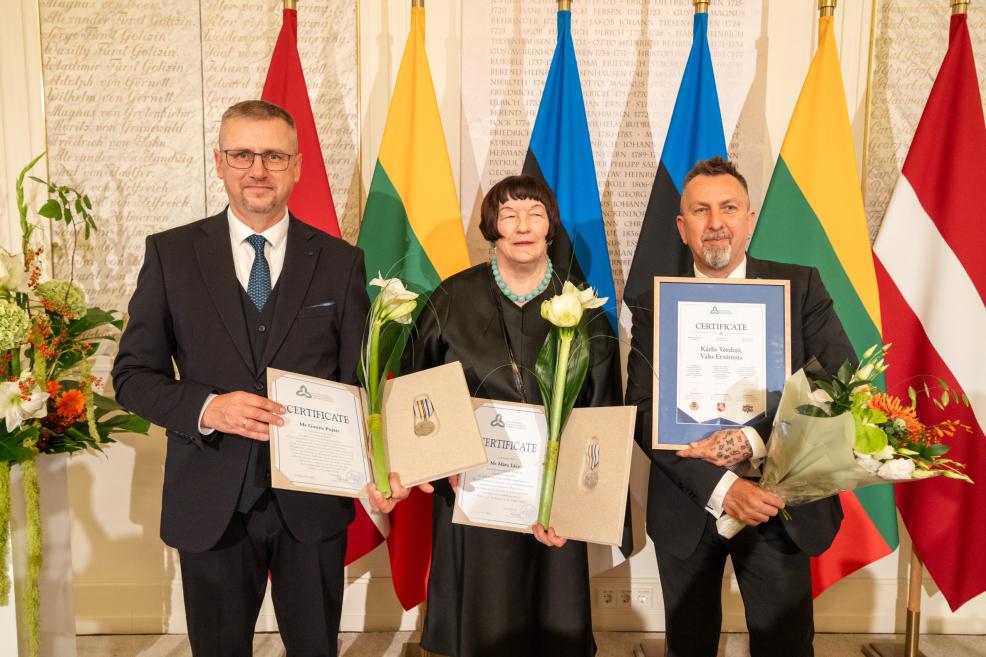 Trīs svinīgi ģērbti cilvēki stāv telpā pie Latvijas karogiem, turot sertifikātus, medaļas un ziedus.