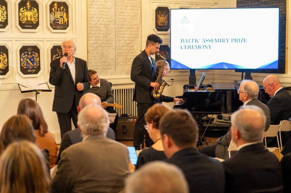 Eleganta zālē notiek Baltijas Asamblejas balvas ceremonija ar runātāju pie mikrofona, saksofonistu un pianistiem klavieru priekšā skatītāju priekšā.