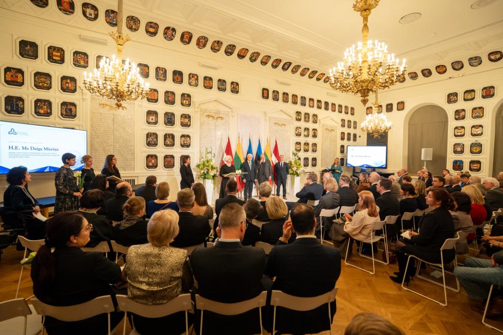 Svinīga pieņemšana lielā zālē ar publiku, kristāla lustrām, Latvijas un citu valstu karogiem pie skatuves un uz ekrāniem redzamu uzrunu H. E. Ms Daiga Mieriņa.