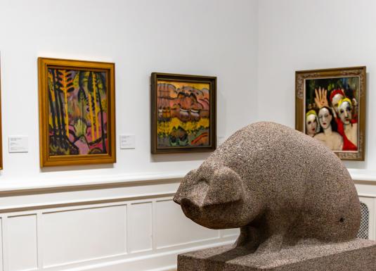 A large stone bear sculpture stands in the foreground of an art gallery room, with several framed paintings hanging on the white walls behind it.