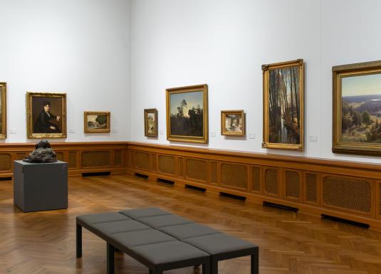 Muzeja galerijas zāle ar gleznām zelta rāmjos uz baltām sienām, koka grīdu, skulptūru uz pjedestāla un polsterētu solu priekšplānā.