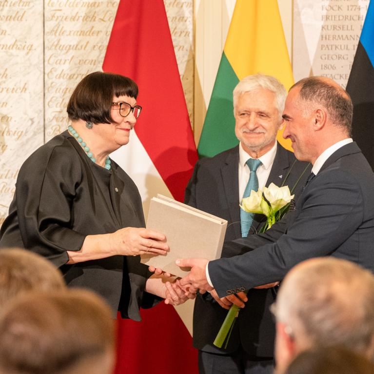 Svinīgā telpā sieviete ar brillēm pasniedz dokumentu uzvalkā ģērbtam vīrietim, fonā stāv vēl divi vīrieši un vairākas valsts karogu joslas.