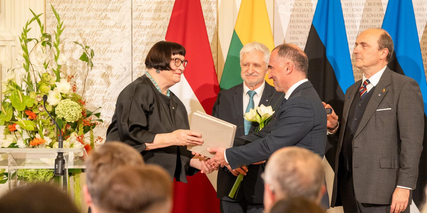 Svinīgā telpā sieviete ar brillēm pasniedz dokumentu uzvalkā ģērbtam vīrietim, fonā stāv vēl divi vīrieši un vairākas valsts karogu joslas.