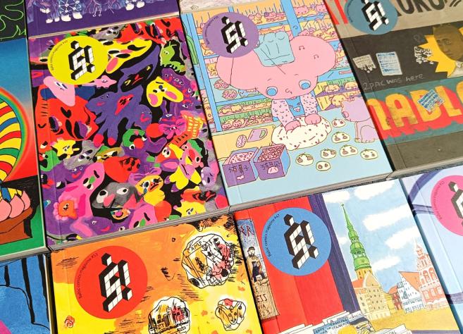 Uz galda izvietoti vairāki krāsaini Baltic Comics Magazine izdevumi ar spilgtiem, dažādiem vāku ilustrāciju motīviem.