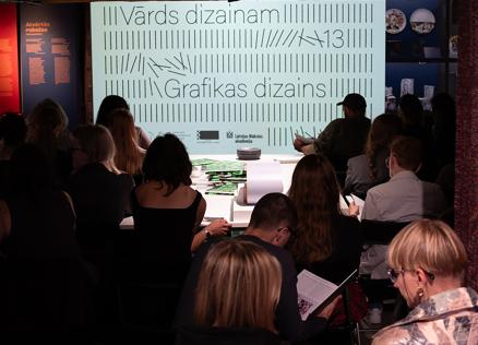 Muzeja zālē no mugurpuses redzami klausītāji, kas sēž auditorijā pie ekrāna ar uzrakstiem Vārds dizainam un Grafikas dizains.