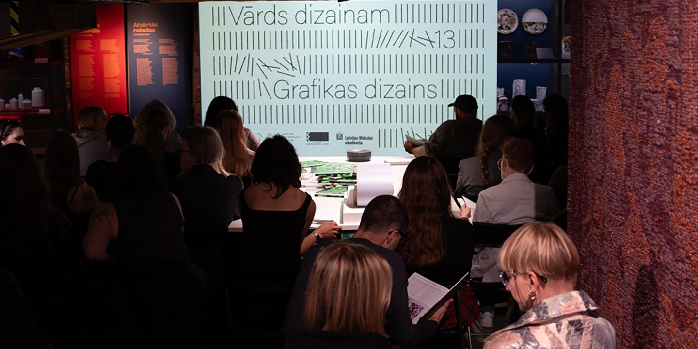 Muzeja zālē no mugurpuses redzami klausītāji, kas sēž auditorijā pie ekrāna ar uzrakstiem Vārds dizainam un Grafikas dizains.