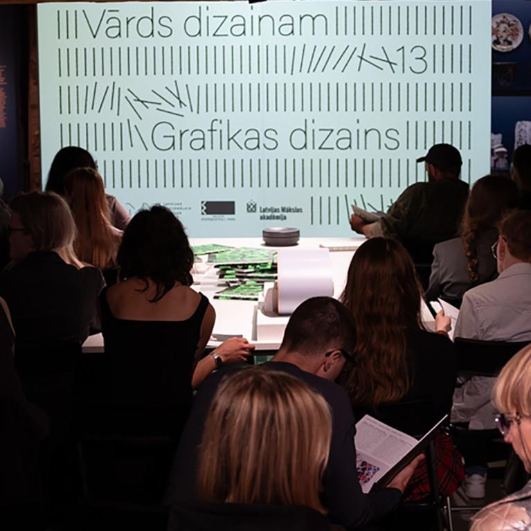 Muzeja zālē no mugurpuses redzami klausītāji, kas sēž auditorijā pie ekrāna ar uzrakstiem Vārds dizainam un Grafikas dizains.