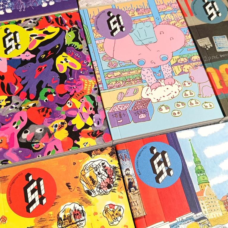 Uz galda izvietoti vairāki krāsaini Baltic Comics Magazine izdevumi ar spilgtiem, dažādiem vāku ilustrāciju motīviem.