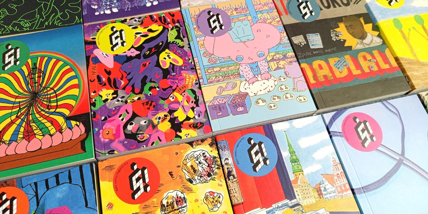Uz galda izvietoti vairāki krāsaini Baltic Comics Magazine izdevumi ar spilgtiem, dažādiem vāku ilustrāciju motīviem.