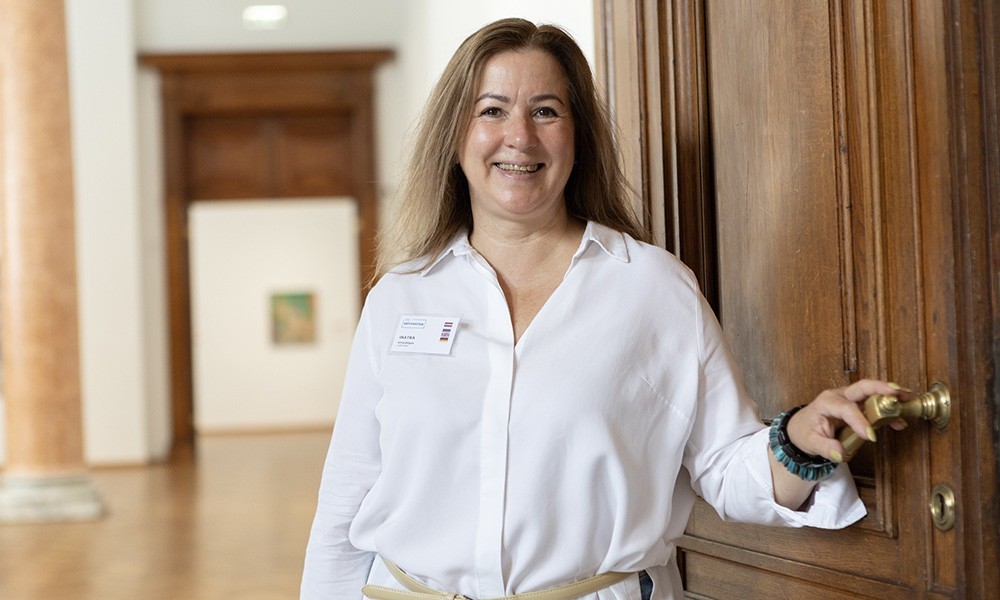 The image shows a smiling museum employee in a white blouse standing by an open wooden door inside the museum.