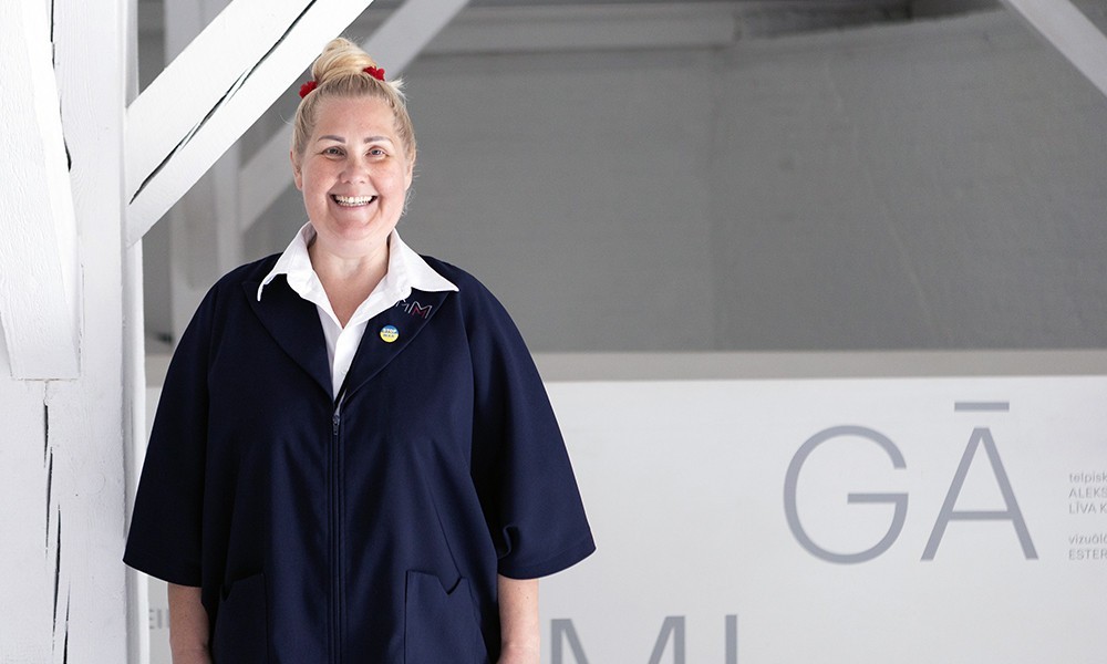 The image shows a smiling museum employee in a dark uniform standing near a staircase inside the museum.