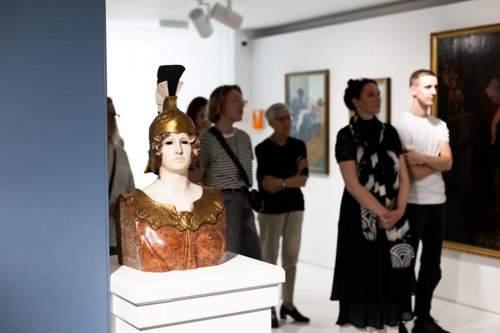 Visitors stand by a wall with paintings at an art exhibition, with a sculpture of a woman in Roman armor on a pedestal in the foreground.