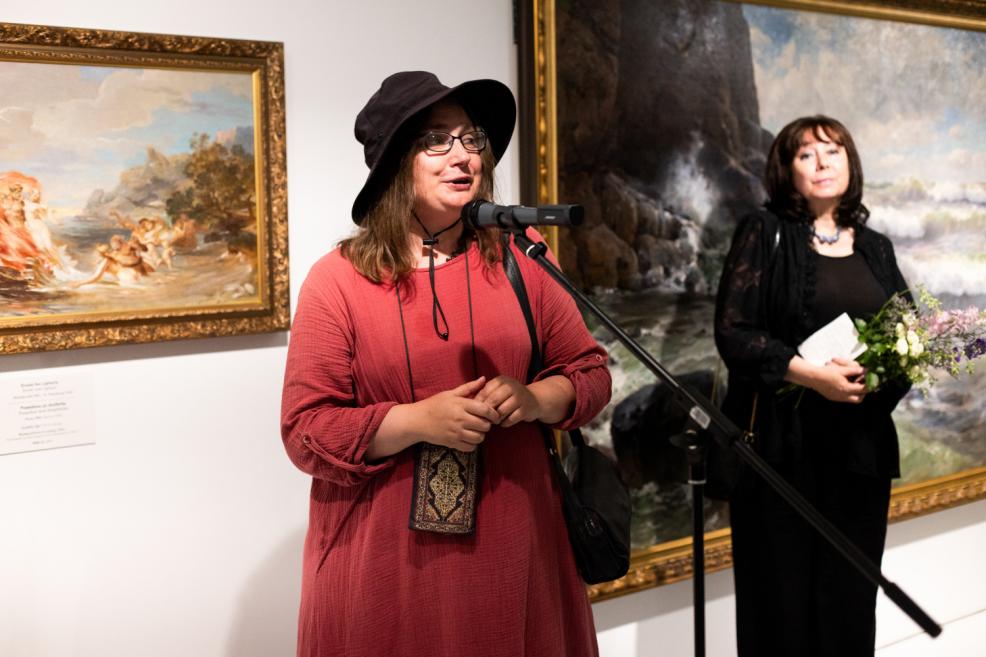A woman wearing a wide-brimmed hat and glasses speaks into a microphone at an art exhibition, with paintings and another woman holding flowers in the background.