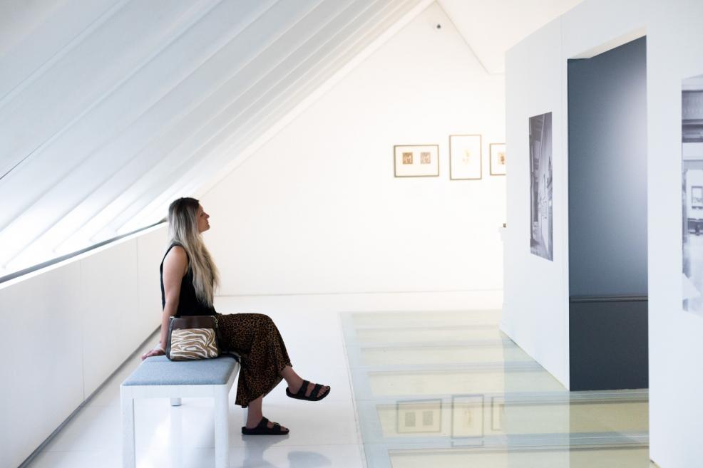 A woman sits on a bench in a white exhibition hall beneath a sloped ceiling window, observing artworks on the wall.