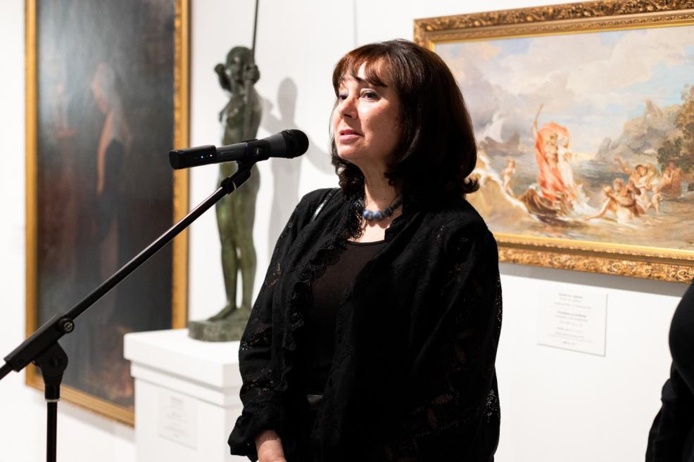 A woman in black speaks into a microphone at an art exhibition opening, with paintings and a sculpture visible in the background.