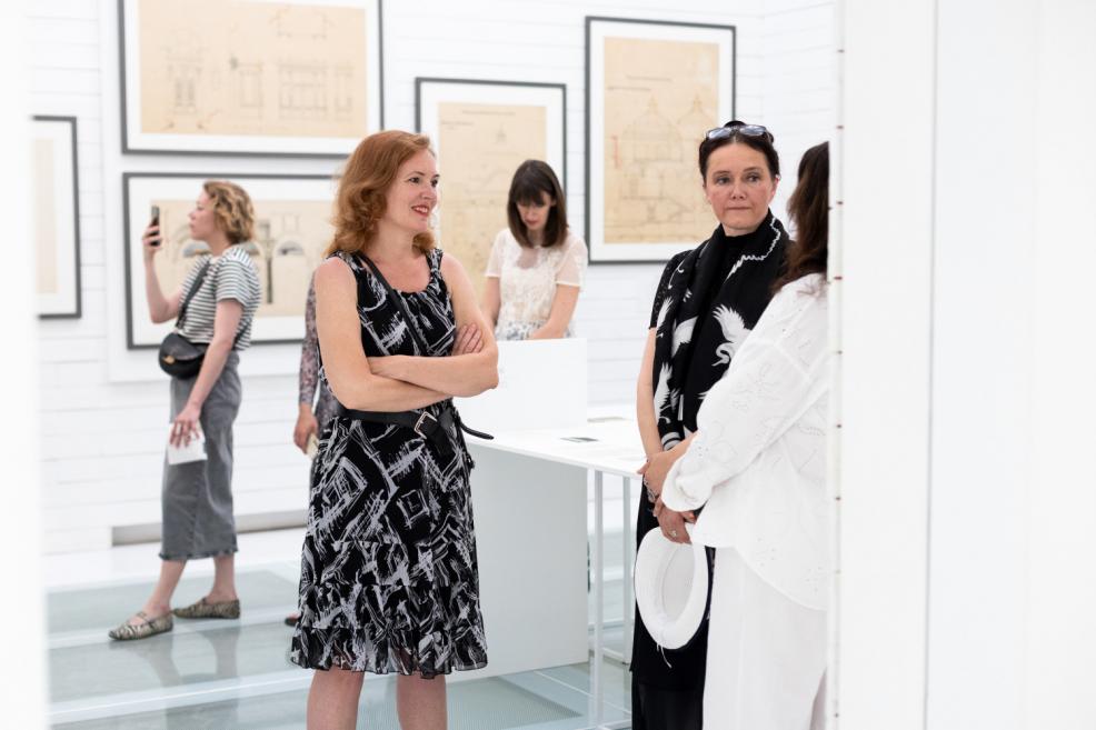 Women conversing and viewing architectural drawings in a white-walled exhibition space.