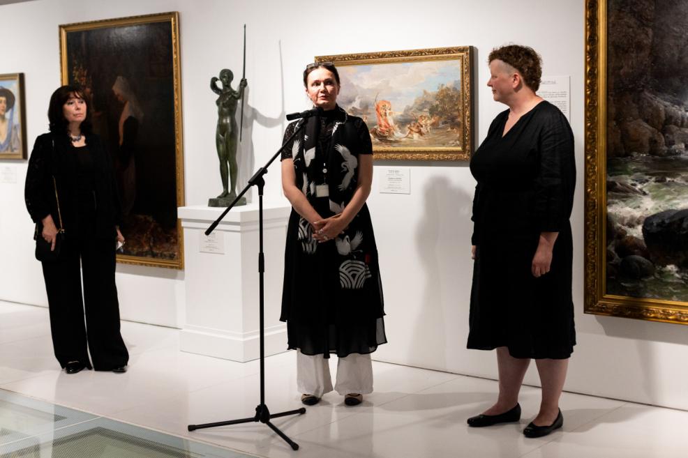 Three women stand near a microphone in an art exhibition hall, with paintings and a bronze sculpture in the background.