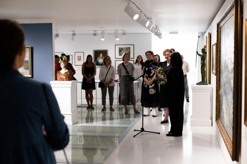 Art exhibition visitors listen to a speaker at the microphone, with paintings and sculptures visible in the hall and a woman holding a bouquet in the foreground.