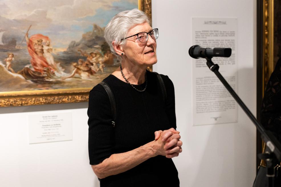A grey-haired woman wearing glasses speaks into a microphone at an art exhibition, with a mythological painting featuring Neptune in the background.