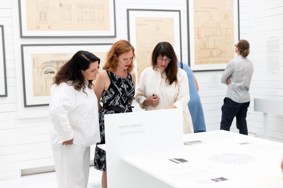 Three women examine exhibition materials featuring architectural drawings in a museum display, with other visitors and sketches visible in the background.