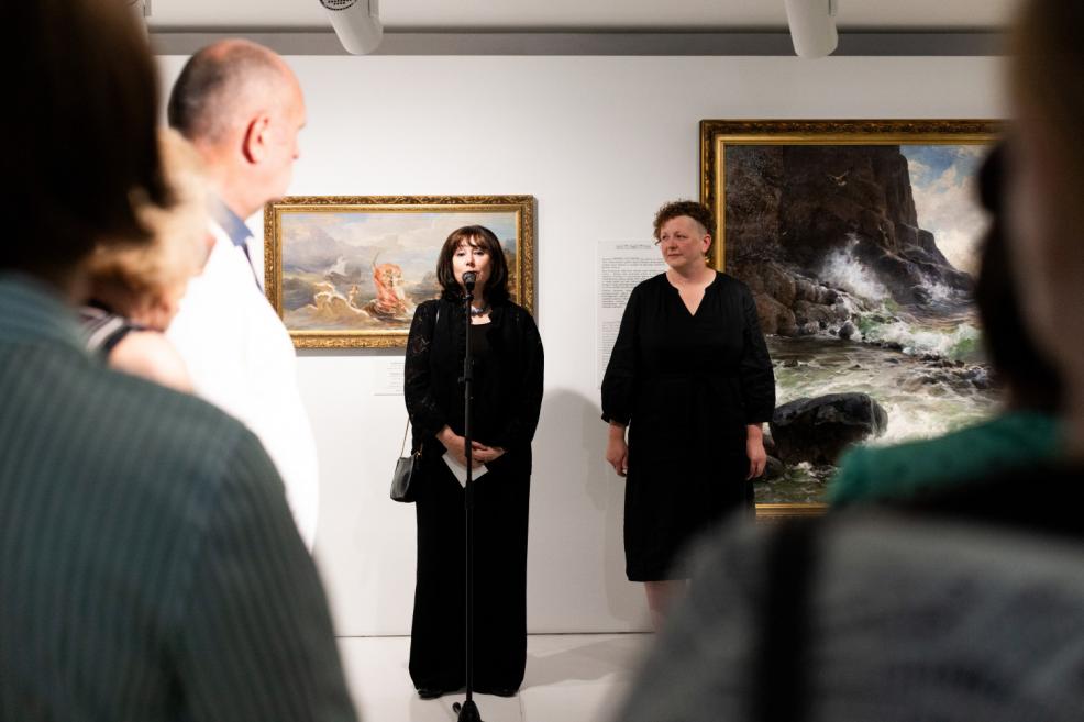 A woman speaks into a microphone at an art exhibition opening, in front of an audience, with seascape paintings in the background.