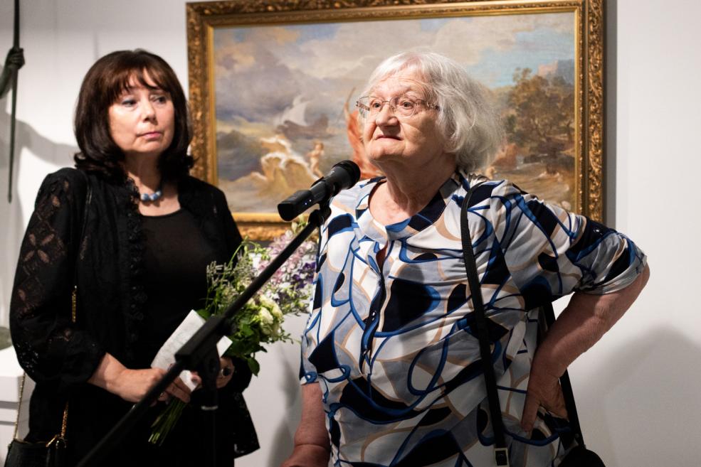 An elderly woman speaks into a microphone at an art exhibition, with another woman holding flowers beside her and a mythological painting in the background.