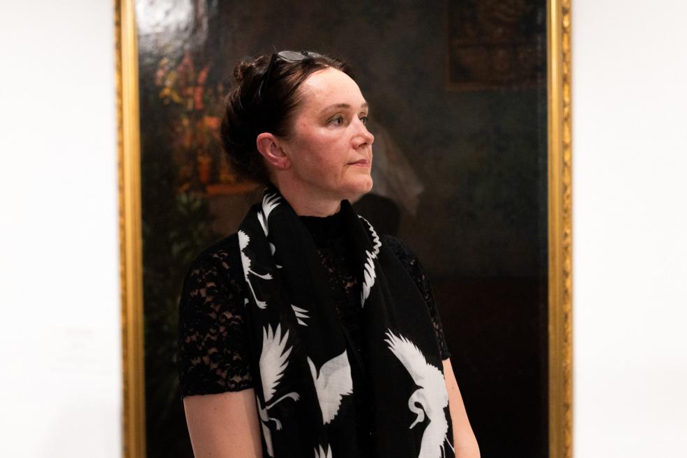 A woman wearing a black scarf with white bird motifs stands in an art exhibition, with a dark-toned painting in the background.