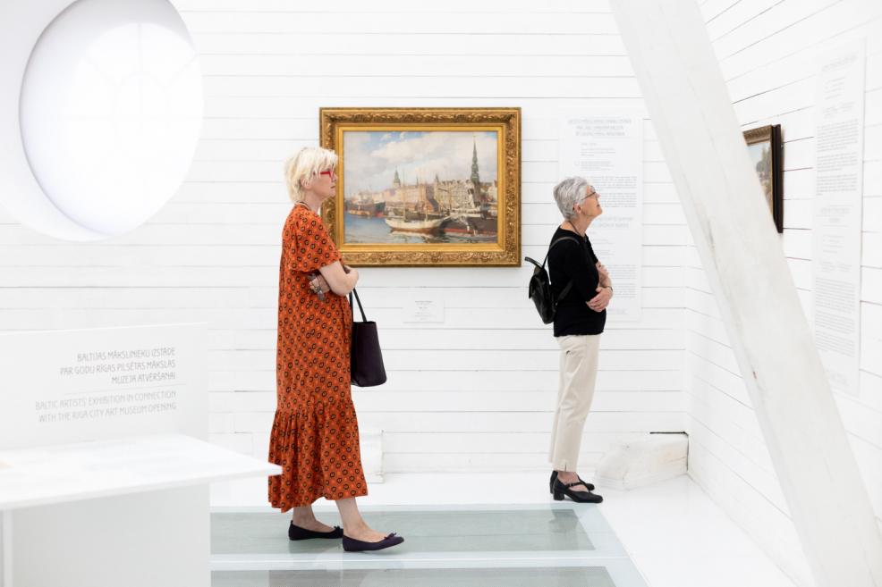 Two women observing a cityscape painting in a bright, white exhibition hall.