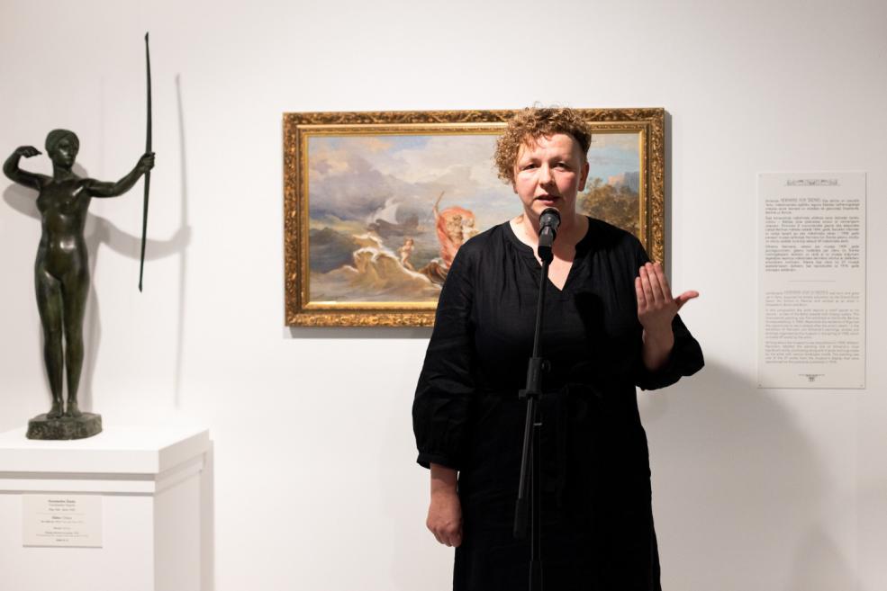 A woman in black speaks into a microphone in an exhibition hall, with a mythological painting and a bronze archer sculpture in the background.