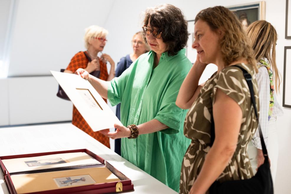 A woman in a green shirt shows an artwork on a cardboard sheet to another visitor in the exhibition hall, with more women visible in the background.