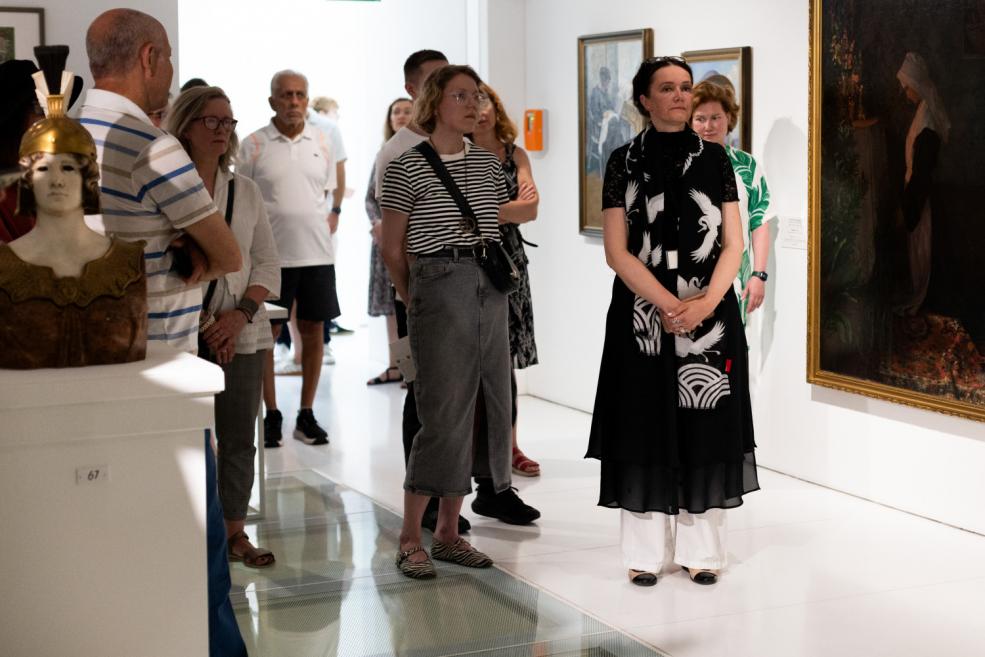Visitors stand and observe a large painting in an art exhibition hall, with a woman in black and white clothing in the foreground.