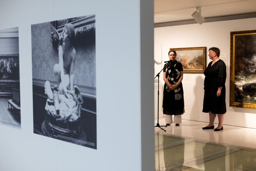 Two women stand near a microphone in an exhibition space with paintings, while black-and-white art photographs are visible in the foreground on the wall.