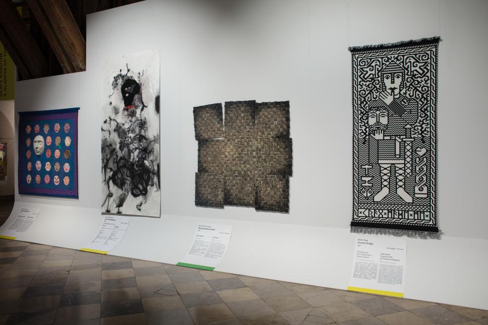 Pie baltas sienas rindā izvietoti četri darbi – daudzkrāsaini sejas motīvi, melnbalts figurāls darbs, tumšs tekstila gabals un melnbalts ornamentiāls paklājs ar cilvēku figūrām.