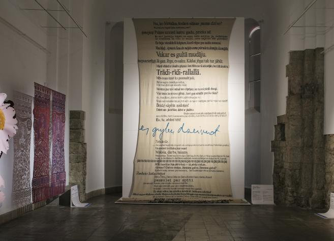 Contemporary art exhibition hall with a three-dimensional floral composition, textile works, a large canvas with inscriptions, and a black canvas with linear figures.