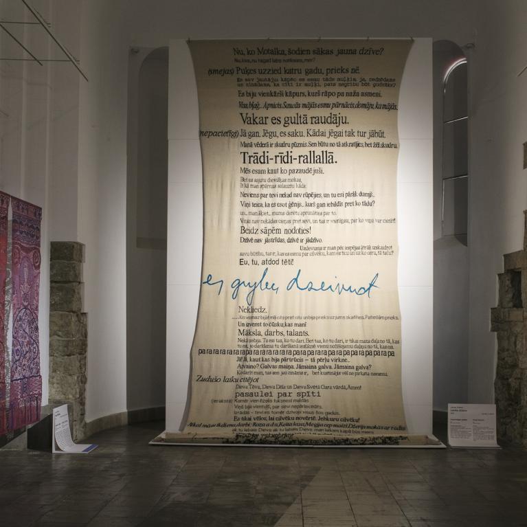 Izstāžu telpa ar lielu audeklu ar latviešu tekstiem centrā, krāsainu telpisku darbu un tekstila paneļiem kreisajā pusē, kā arī melnbaltu gleznojumu uz akmens sienas labajā pusē.