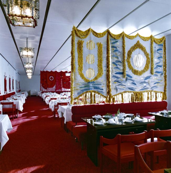 Restaurant interior with a red carpet, tables laid with white cloths, and large decorative textile wall hangings.