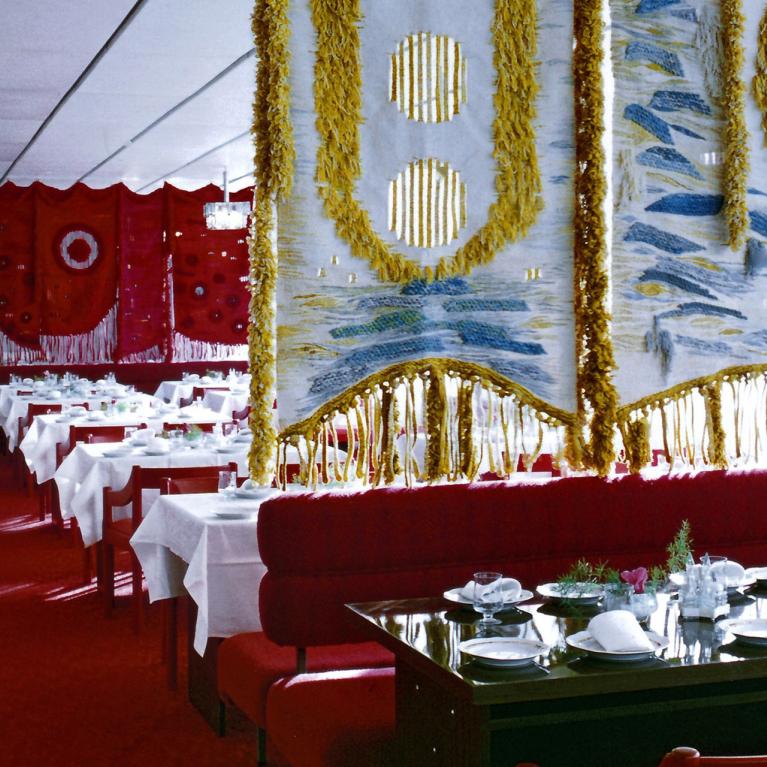 Restaurant dining room with red furniture, white tablecloths and large decorative textiles on the walls.