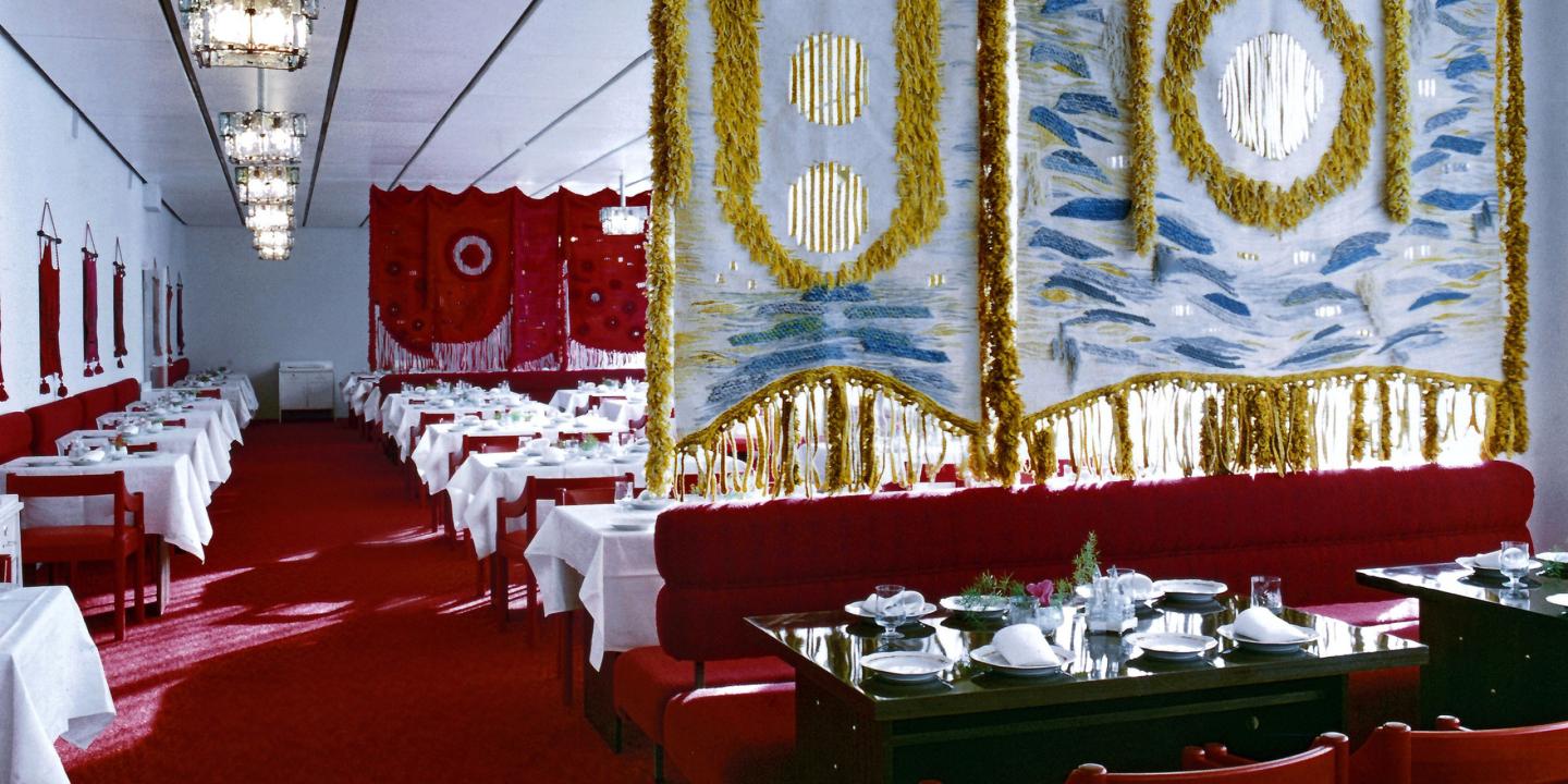 Restaurant dining room with red furniture, white tablecloths and large decorative textiles on the walls.