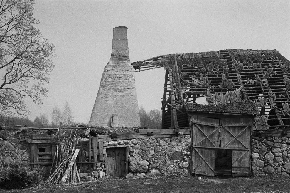 Melnbalta fotogrāfija ar pamestu akmens un koka ēku ar sabrukušu jumtu un augstu skursteni, priekšplānā koka vārti un akmens mūris.