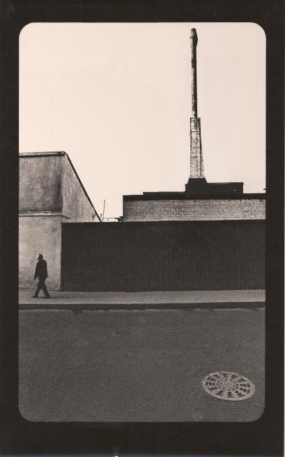 Melnbalta fotogrāfija ar industriālu ēku fasādēm, augstu skursteni un ejā pa kreisi ejošu cilvēka siluetu.