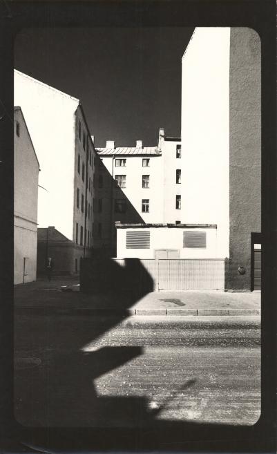 Melnbalta fotogrāfija ar šauru pilsētas pagalmu starp augstām ēkām, ko šķeļ asas gaismas un ēnu laukumi.