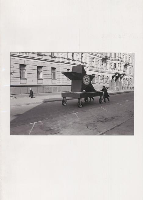 Melnbaltā ielu fotogrāfijā divi cilvēki pārvieto lielu zvaigznes formas objektu ar riteņiem gar pilsētas ielu pie vēsturiskas ēkas.